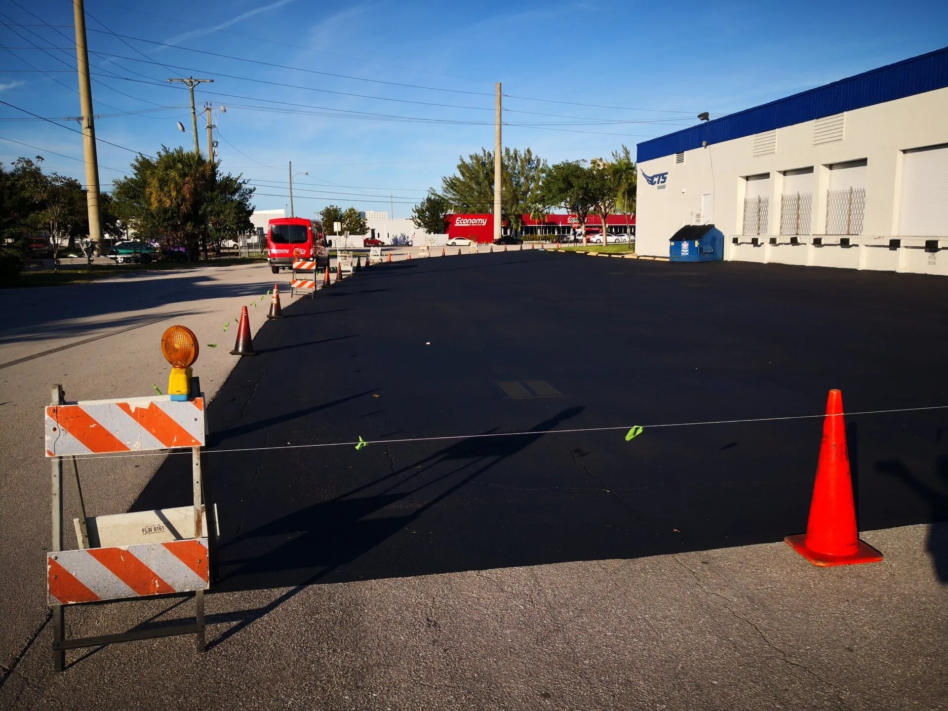 A recently paved parking lot by Jacksonville Asphalt is cordoned off with traffic cones and a barricade to keep vehicles off the new asphalt. An orange warning light is attached to the barricade. In the background, there are trees, power lines, and buildings with colorful signs.